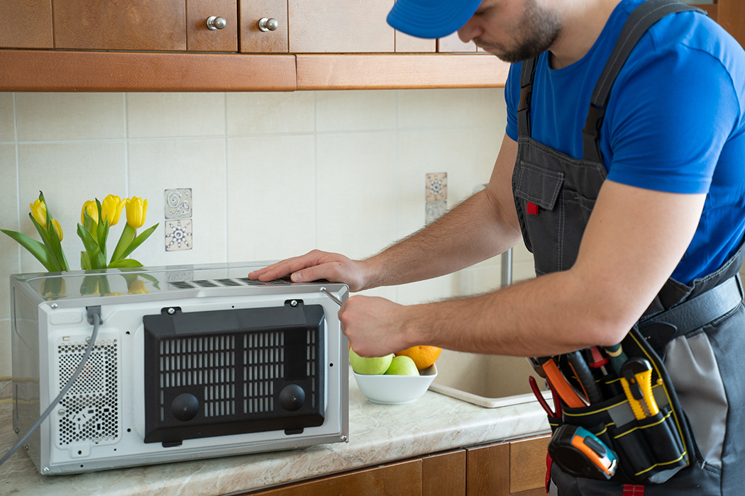 Young repairman fixing and repairing microwave oven by screwdriver in kitchen.